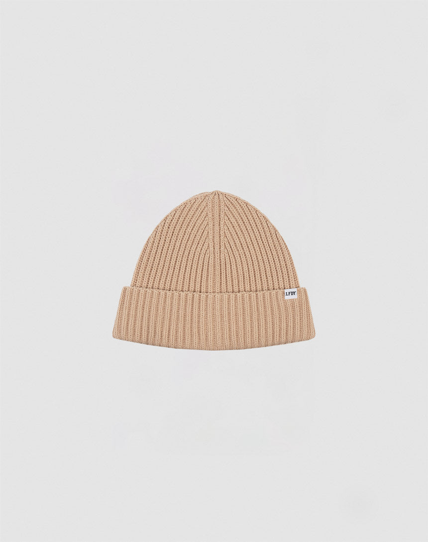 A beige Washed Rib Knit Beanie von LIVE FAST DIE YOUNG aus Baumwolle mit umgeschlagenem Rand und kleinem weißem Etikett an der Seite vor schlicht hellgrauem Hintergrund.