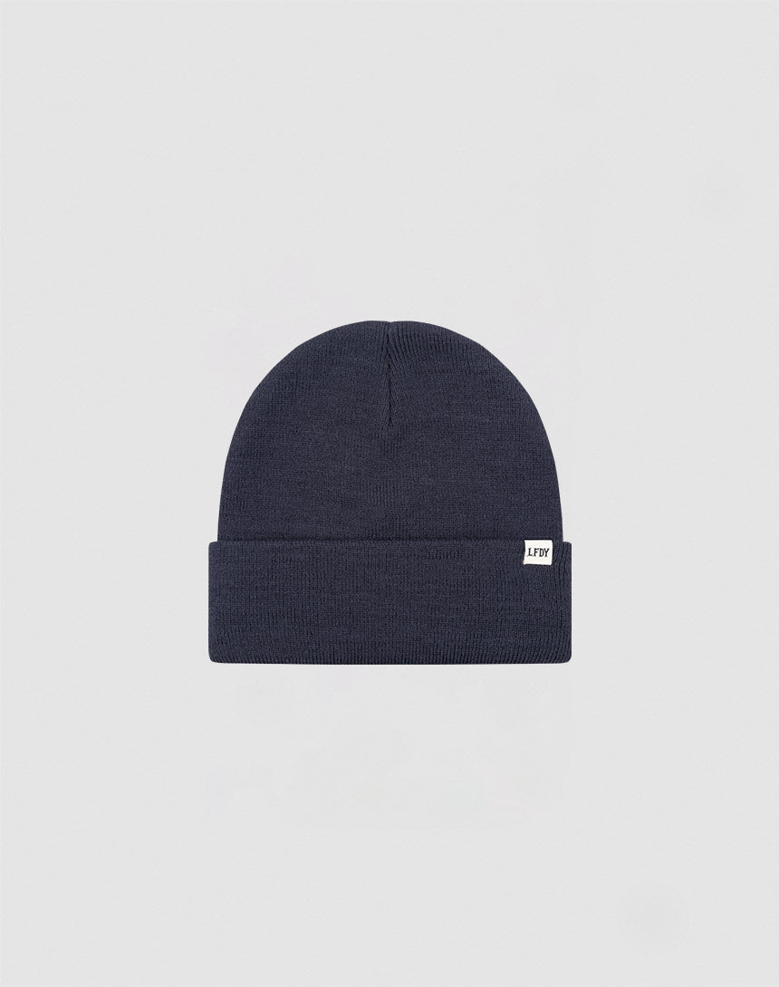 The LIVE FAST DIE YOUNG Basic Beanie is a plain dark navy blue knit, One Size, with a folded brim and a small white Flaglabel tag on the front, displayed against a light gray background.
