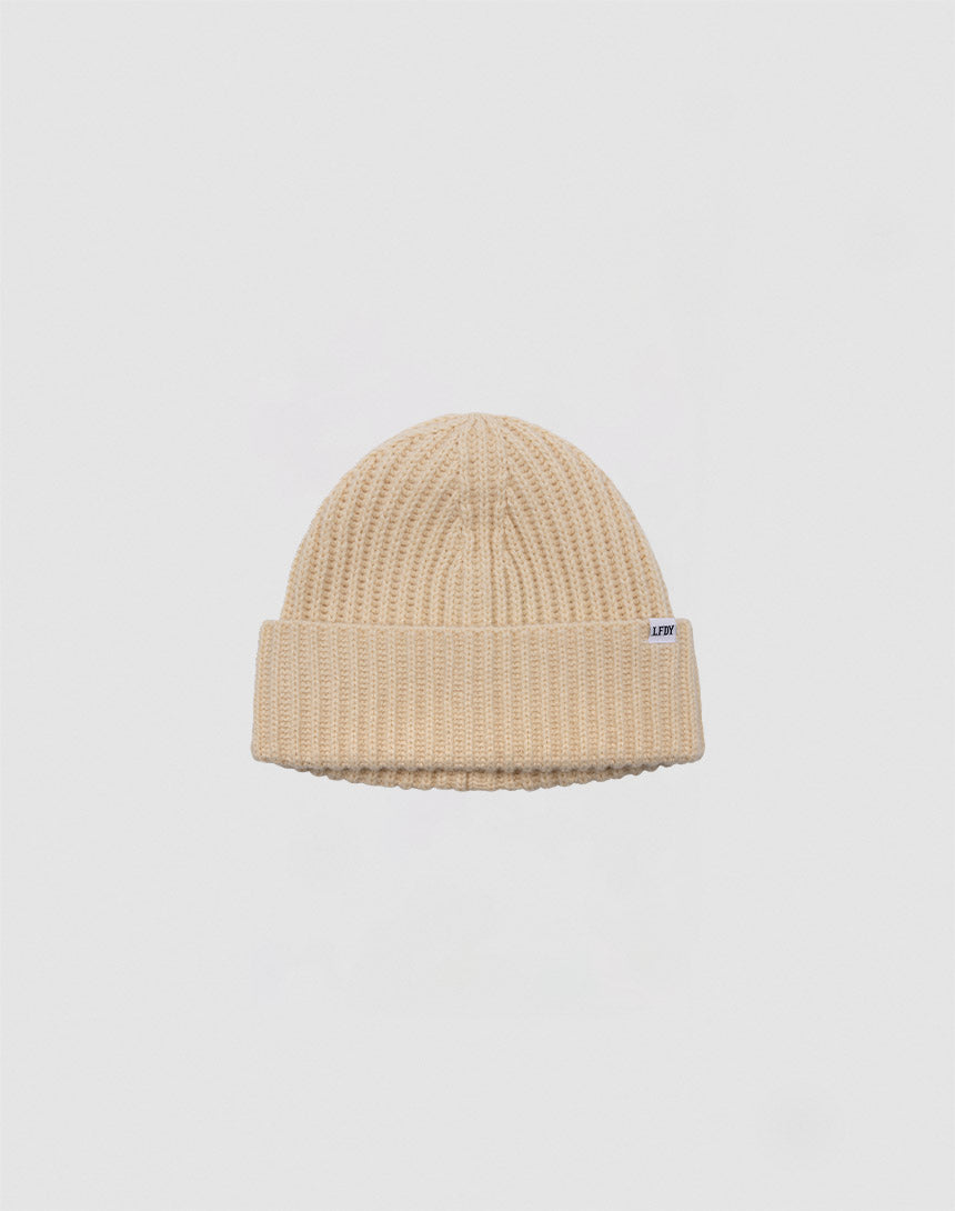 Die Rib Knit Beanie von LFDY in Beige besteht aus 100% Acryl, hat einen gefalteten Bund und ein kleines weißes Rechteck-Label an der Seite – präsentiert vor einem schlichten hellgrauen Hintergrund.