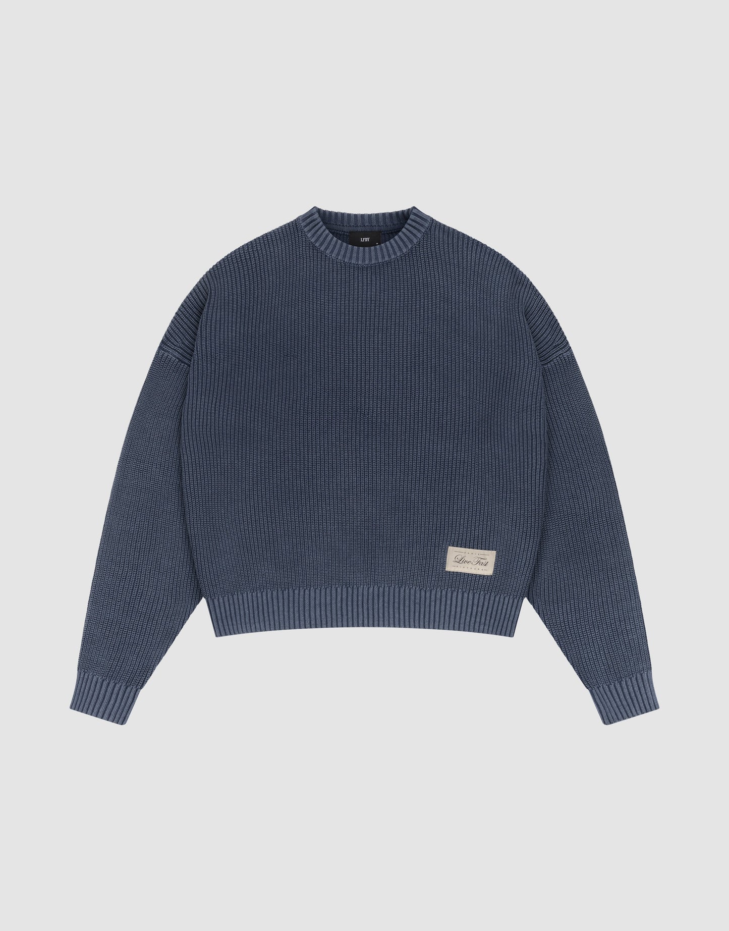 The LFDY Knit Sweater by LIVE FAST DIE YOUNG is a blue ribbed cotton piece with long sleeves, a crew neck, dropped shoulders, an oversized fit, and a beige tag near the hem. It’s shown laid flat on a plain white background.