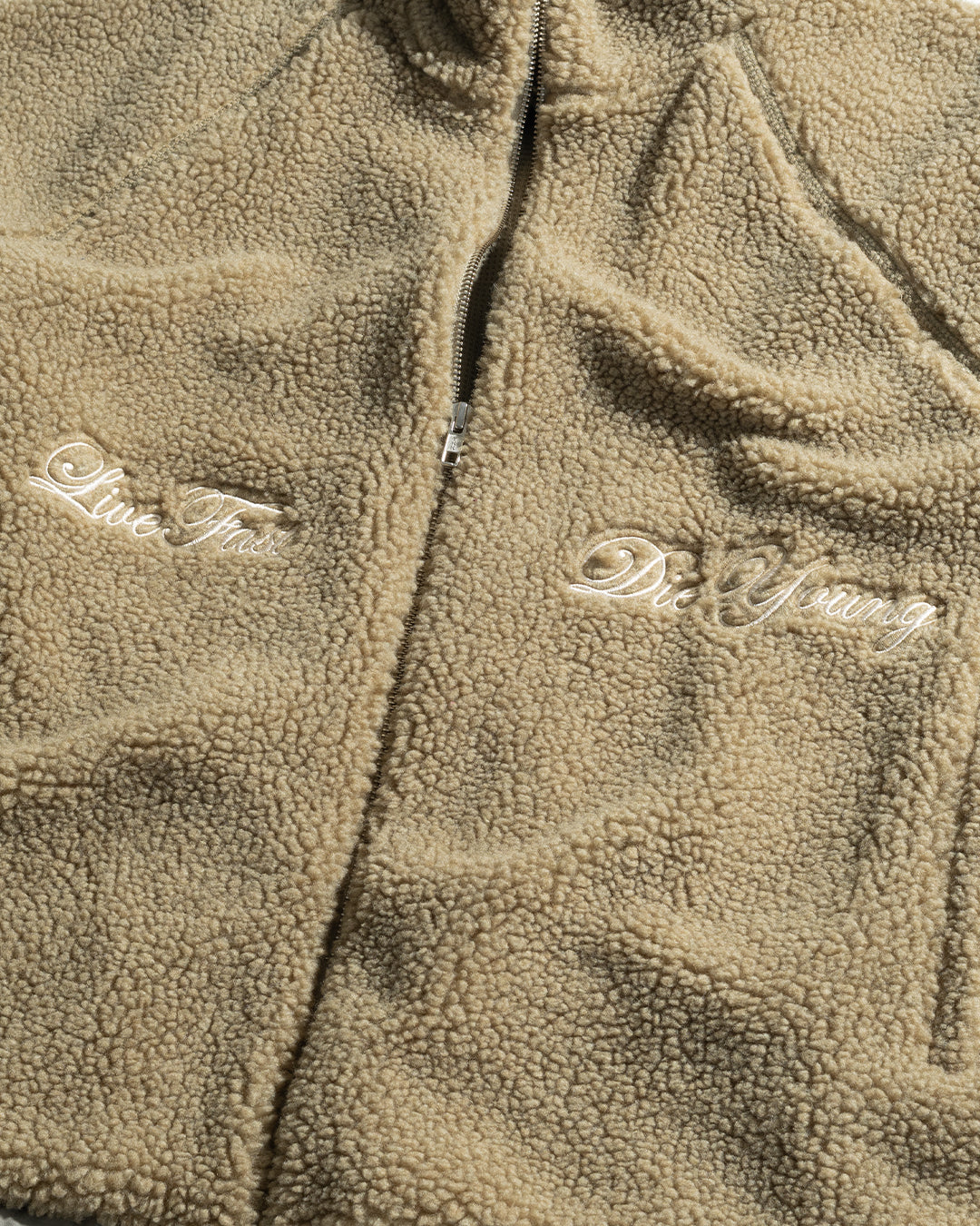 A close-up of the LFDY Teddy Jacket by LIVE FAST DIE YOUNG, featuring a tan color, zipper closure, and white cursive embroidery: “Live Fast” on the left and “Die Young” on the right.