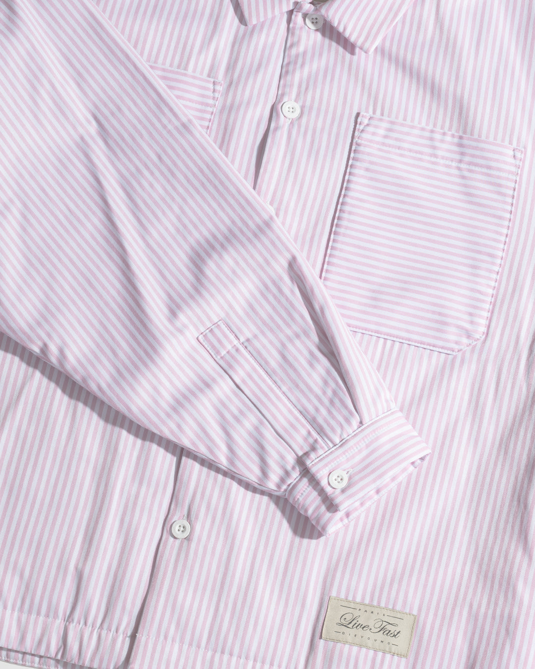 A close-up of the LFDY Striped Padded Shirt by LIVE FAST DIE YOUNG, featuring pink and white vertical stripes, a front pocket, visible cuff, and a small fabric label near the hem that emphasizes its trendy overshirt style.