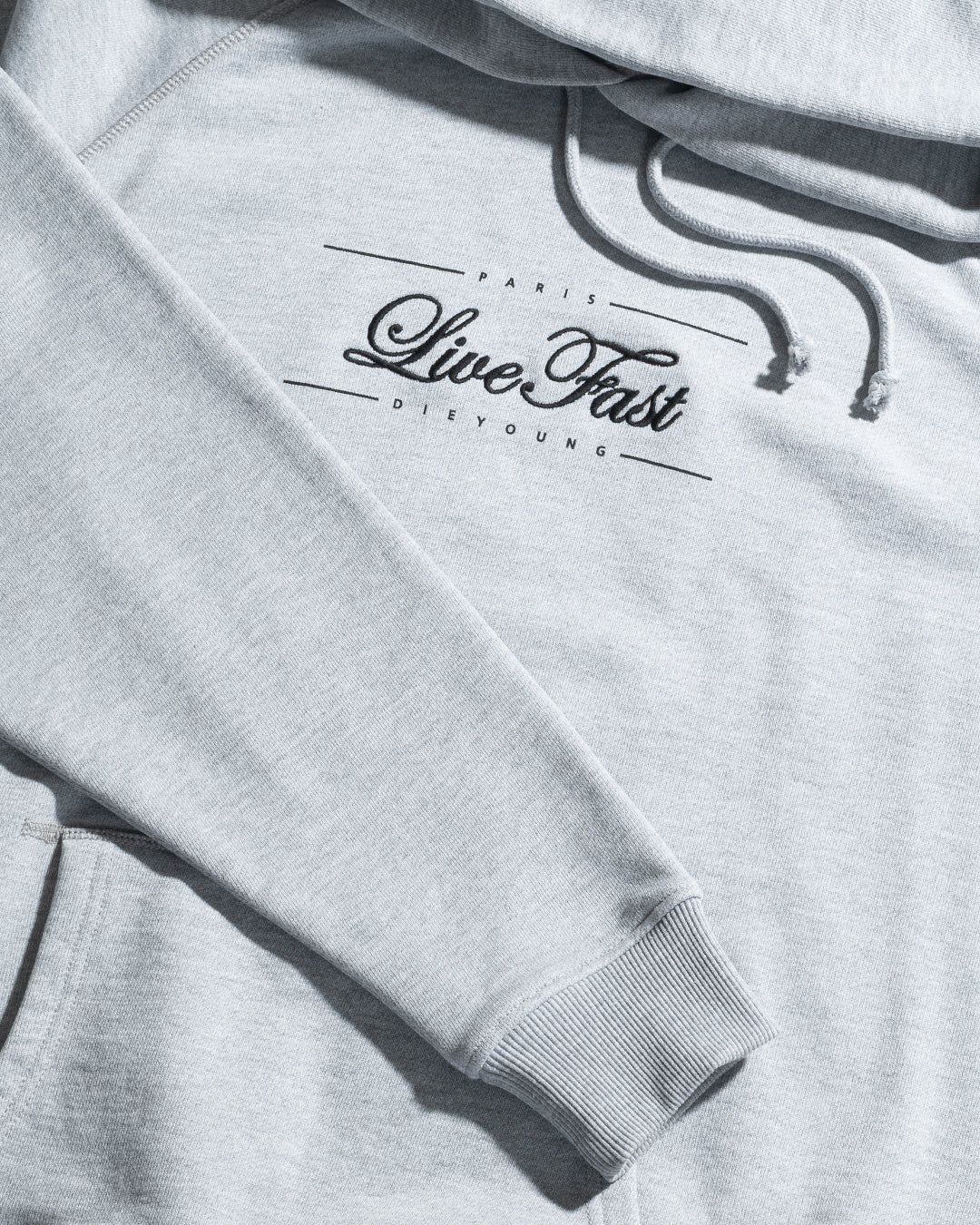 The LFDY Embroidery Hooded by LIVE FAST DIE YOUNG is a light gray regular fit hoodie with raglan sleeves, an adjustable hood, front pocket, ribbed cuffs, and black "Paris Live Fast Die Young" text across the chest.