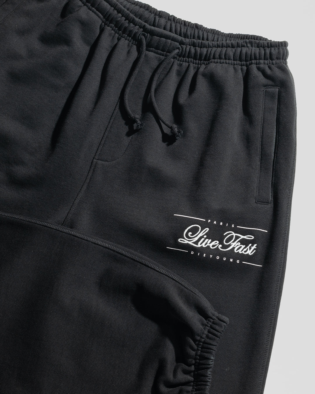 The Wide Cuff Jogger by LIVE FAST DIE YOUNG features an elastic waistband with drawstrings and white script—PARIS Live Fast DIE YOUNG—above the left thigh pocket. Made of soft French Terry, shown on a light gray background.
