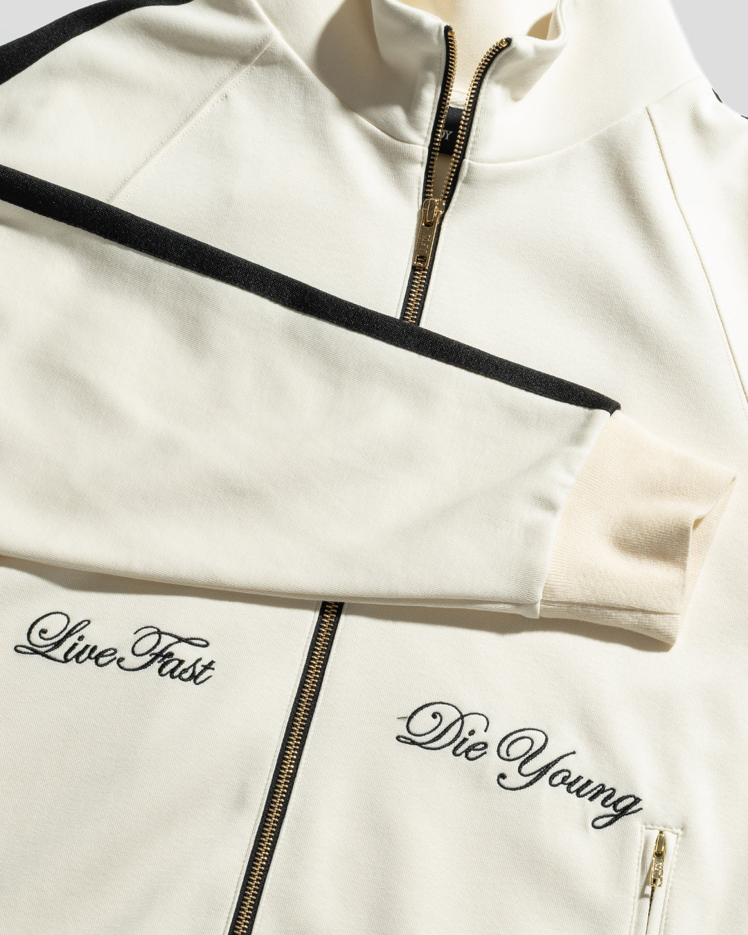 The LFDY Striped Track Jacket by LIVE FAST DIE YOUNG features a cream color with raglan sleeves, black accents, and "Live Fast" and "Die Young" embroidered in black cursive on either side of the zipper.
