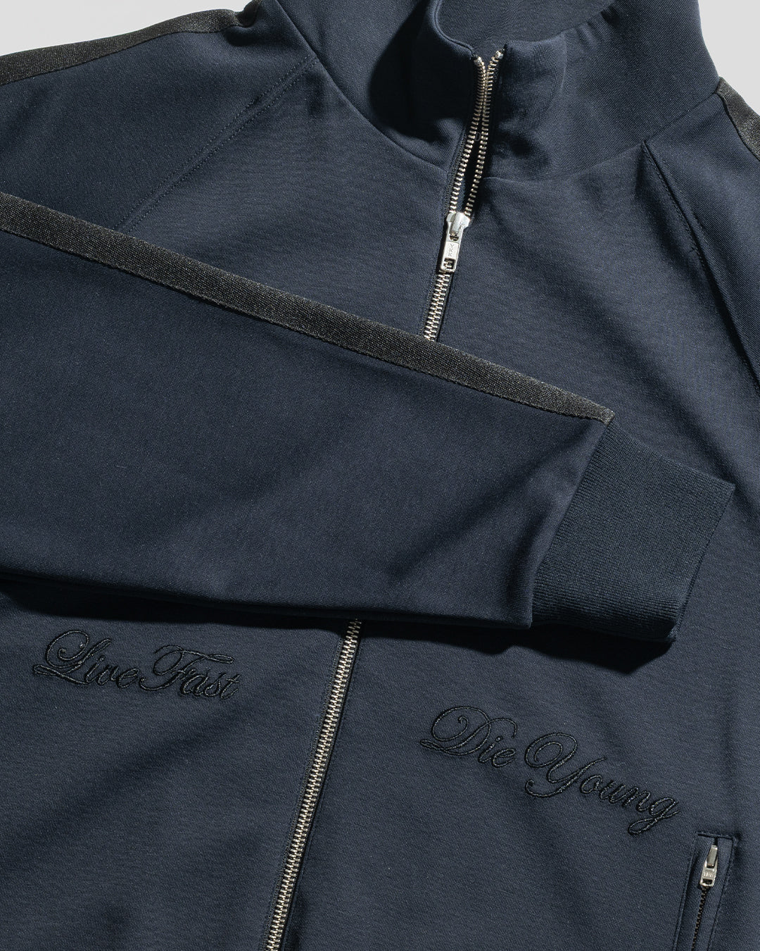 The LFDY Striped Track Jacket by LIVE FAST DIE YOUNG is a dark navy zip-up with black "Live Fast and Die Young" embroidery, high collar, raglan sleeves, ribbed cuffs, and herringbone tape details. Shown against a light grey background.