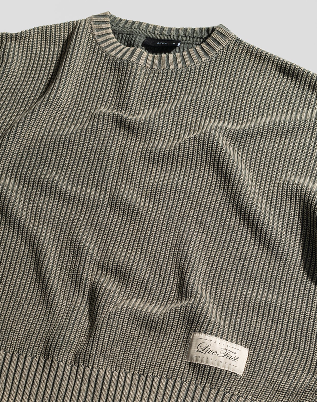The LFDY Knit Sweater by LIVE FAST DIE YOUNG is a beige and gray ribbed, oversized pullover with a round neck, dropped shoulders, and a “Live Fast” patch near the hem. Made from 100% Baumwolle, shown laid flat on a light surface.