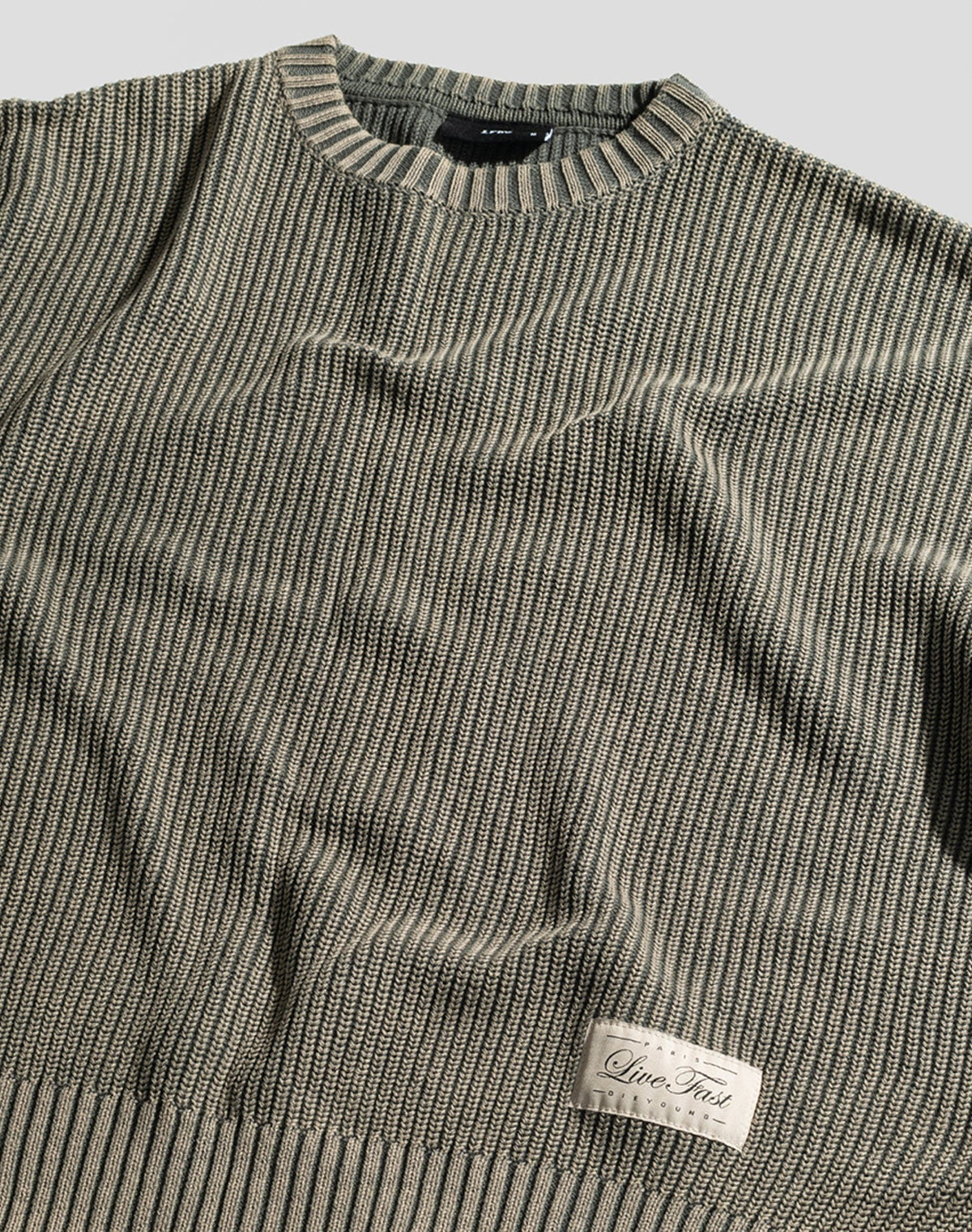 The LFDY Knit Sweater by LIVE FAST DIE YOUNG is a beige and gray ribbed, oversized pullover with a round neck, dropped shoulders, and a “Live Fast” patch near the hem. Made from 100% Baumwolle, shown laid flat on a light surface.
