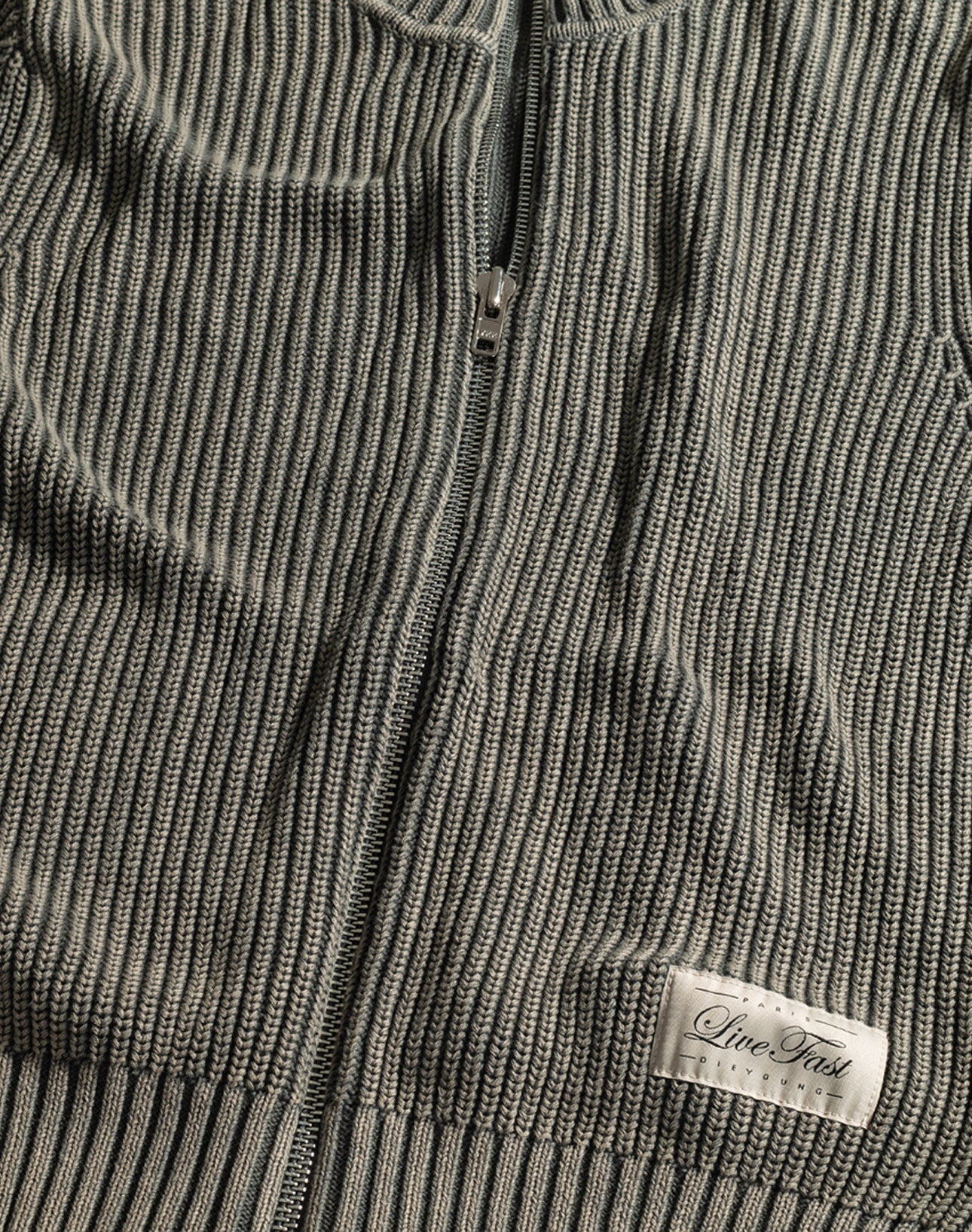 Close-up of the LFDY Knit Jacket by LIVE FAST DIE YOUNG—a ribbed gray zip-up with a stand collar and a small white "Live Fast PREMIUM" label near the hem. The zipper is partially visible at the center of this Regular Fit piece.