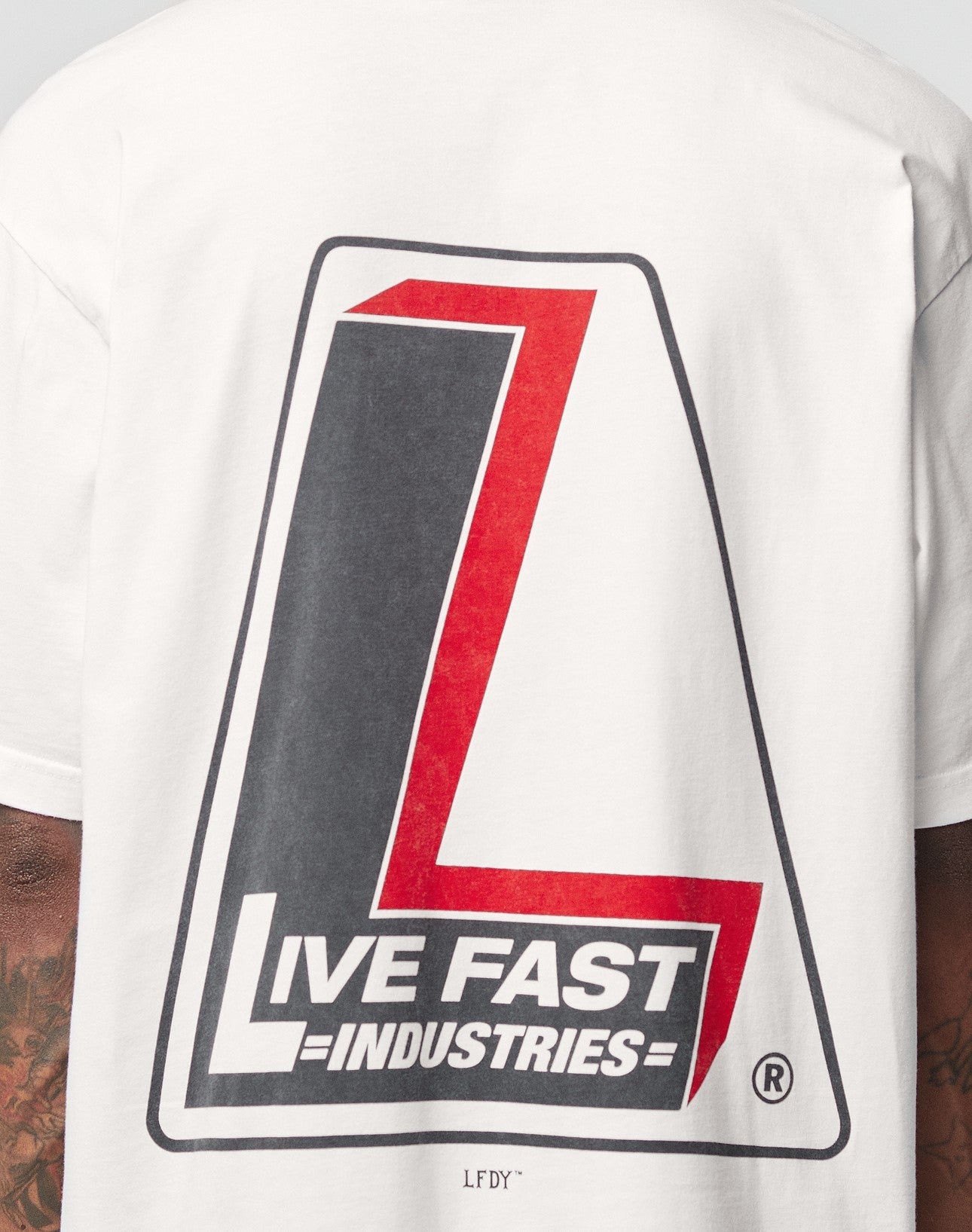 A person wears the LFDY Industries Tee by LIVE FAST DIE YOUNG, an oversize white cotton shirt with bold black and red L and a LIVE FAST INDUSTRIES backprint. Distinct front and back prints, with tattoos visible on their arms.