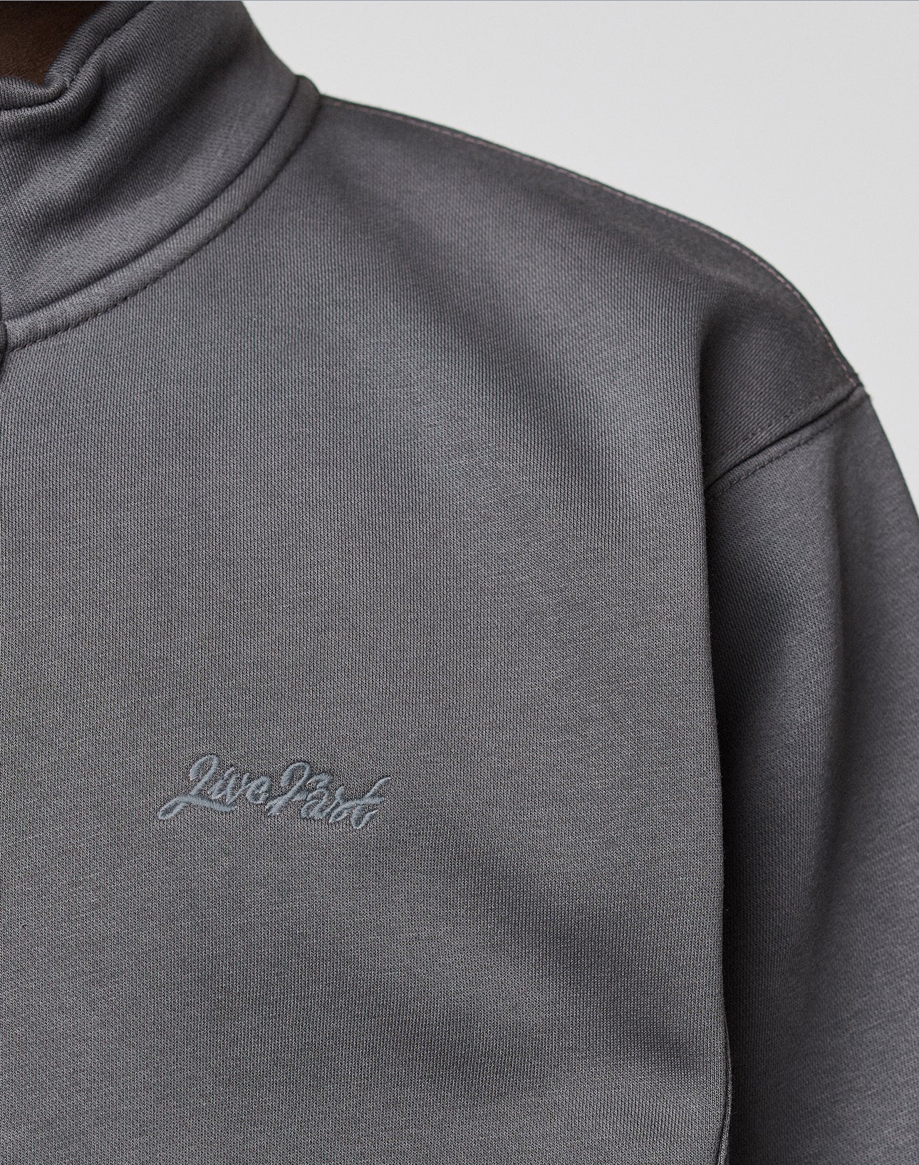 Close-up of a person wearing a grey LIVE FAST DIE YOUNG Sweatsuit Jacket with a standing collar and "Live First" embroidered on the chest in grey thread.