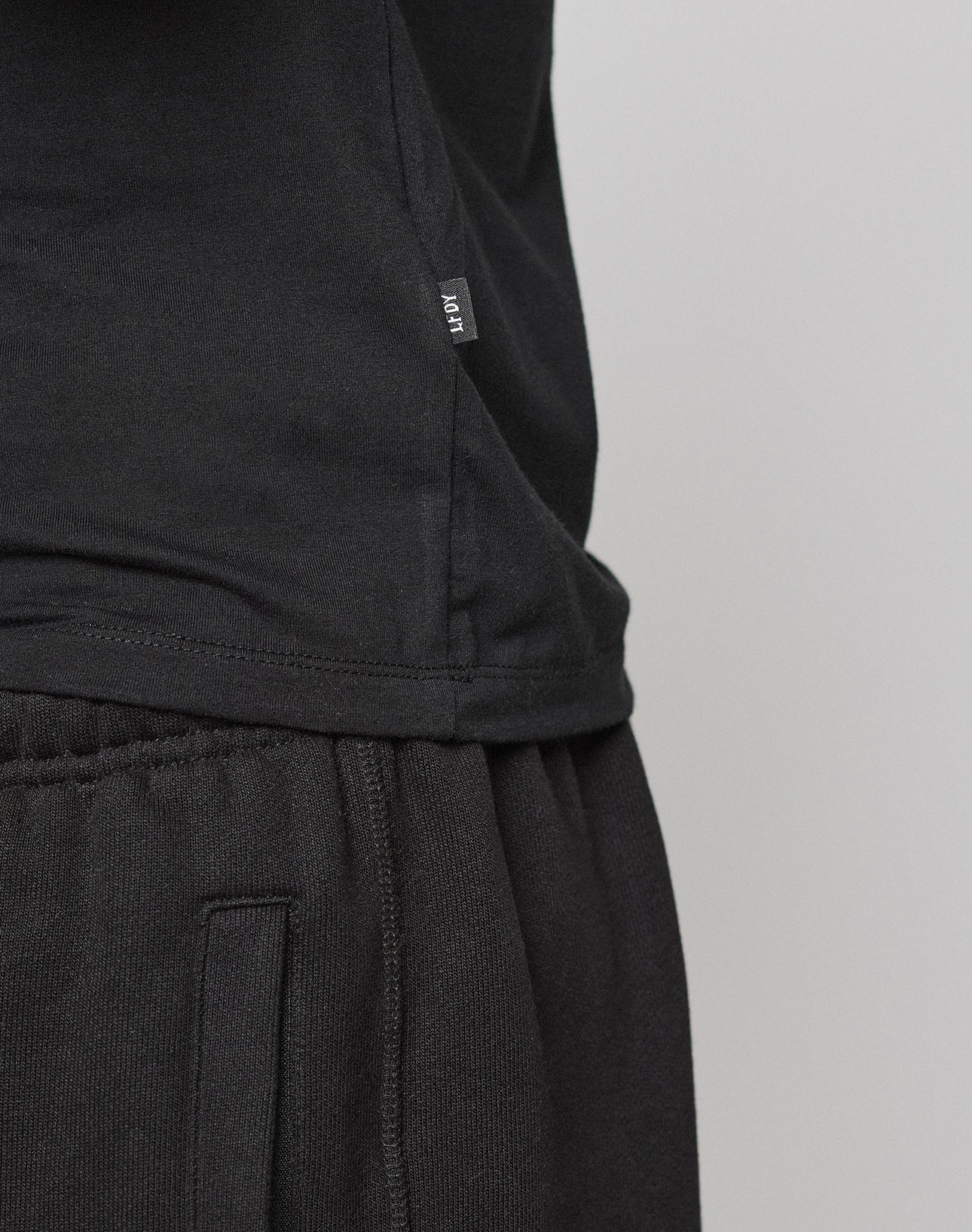 Close-up of a person in a black LFDY Slim Tee by LIVE FAST DIE YOUNG and black sweatpants. The side hem shows a small Flag-Label with white MOVT text. Made from soft Baumwolle, the tee is set against a plain light gray background.