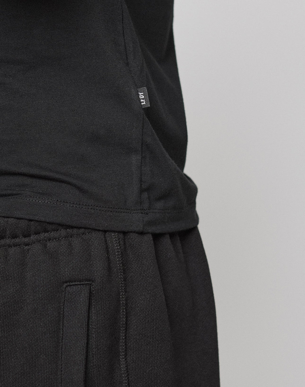 Close-up of a person in a black LFDY Slim Tee by LIVE FAST DIE YOUNG and black sweatpants. The side hem shows a small Flag-Label with white MOVT text. Made from soft Baumwolle, the tee is set against a plain light gray background.