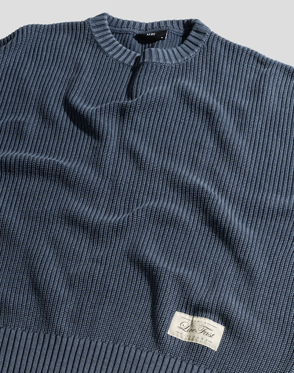 The LFDY Knit Sweater by LIVE FAST DIE YOUNG is shown laid flat in blue ribbed knit with a crew neck, dropped shoulders, and a rectangular white patch with cursive near the hem. The 100% Baumwolle fabric looks soft and lightly wrinkled.