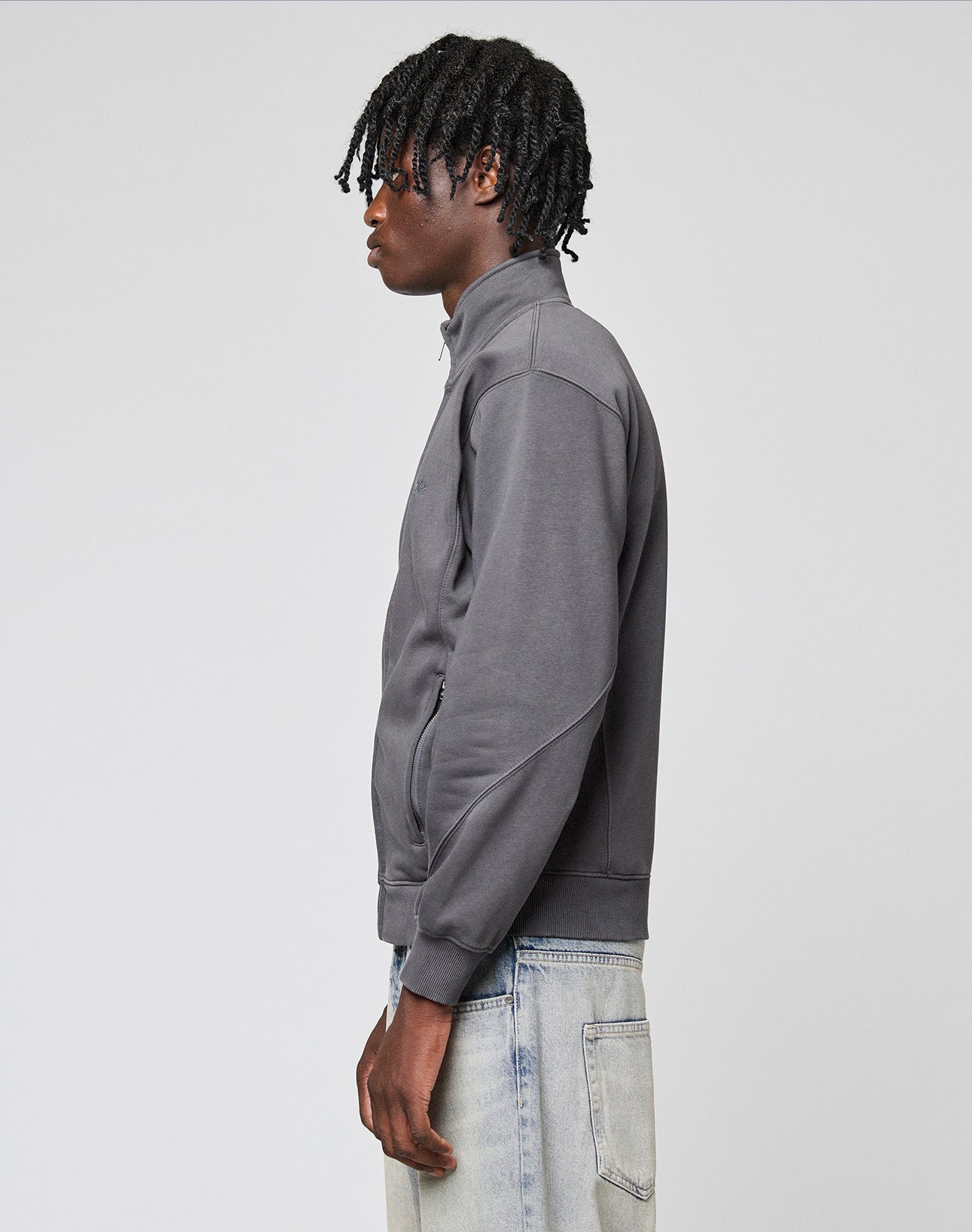 A person with short, twisted hair wears the LIVE FAST DIE YOUNG Sweatsuit Jacket in gray with a Stehkragen and light blue jeans, standing in profile against a plain light gray background.