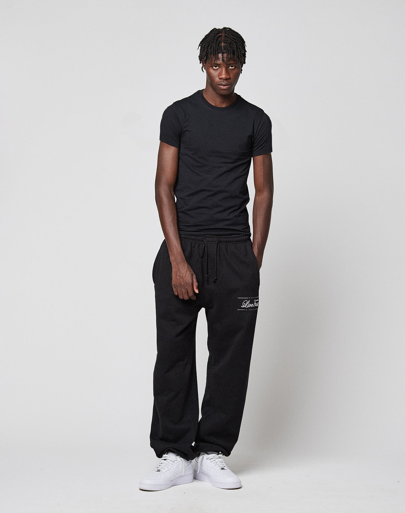 A young man stands against a plain backdrop, wearing the LFDY Slim Tee by LIVE FAST DIE YOUNG, loose black sweatpants with white thigh text and Flag-Label, and white sneakers. His hands are relaxed by his sides.