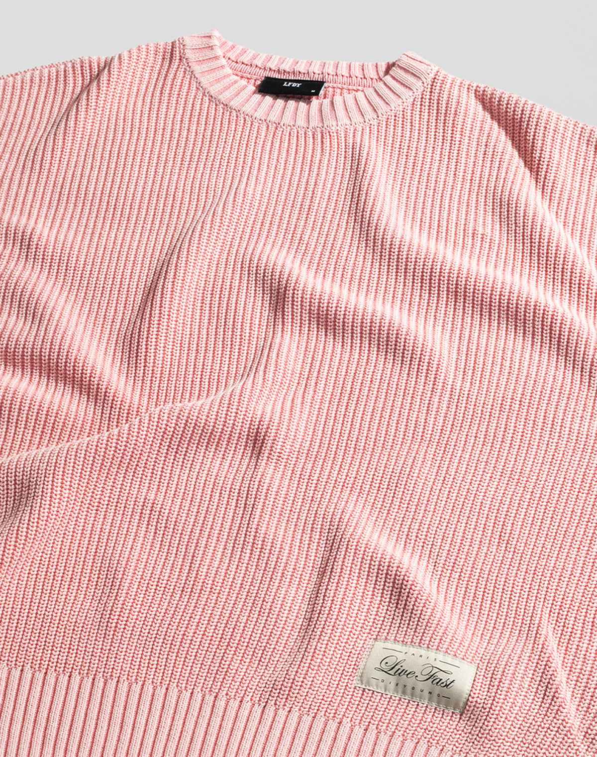 The LFDY Knit Sweater by LIVE FAST DIE YOUNG is shown laid flat in pink and white with a ribbed texture, dropped shoulders, and a small beige "Live Fast" patch near the hem. The neck tag is also visible.