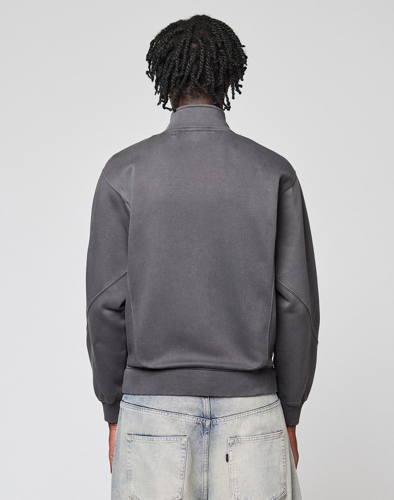 A person with medium-length, twisted hair is seen from behind in a gray LIVE FAST DIE YOUNG Sweatsuit Jacket and light-washed jeans, standing against a plain light gray background.