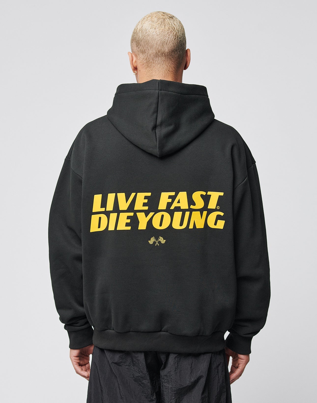 A person with short blond hair wears the LFDY Drift Hooded by LIVE FAST DIE YOUNG, a black hoodie with yellow LIVE FAST. DIE YOUNG text on the back, Oversize Fit, and doppellagige Kapuze, against a plain light background.