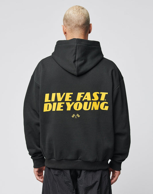 A person with short blond hair wears the LFDY Drift Hooded by LIVE FAST DIE YOUNG, a black hoodie with yellow LIVE FAST. DIE YOUNG text on the back, Oversize Fit, and doppellagige Kapuze, against a plain light background.