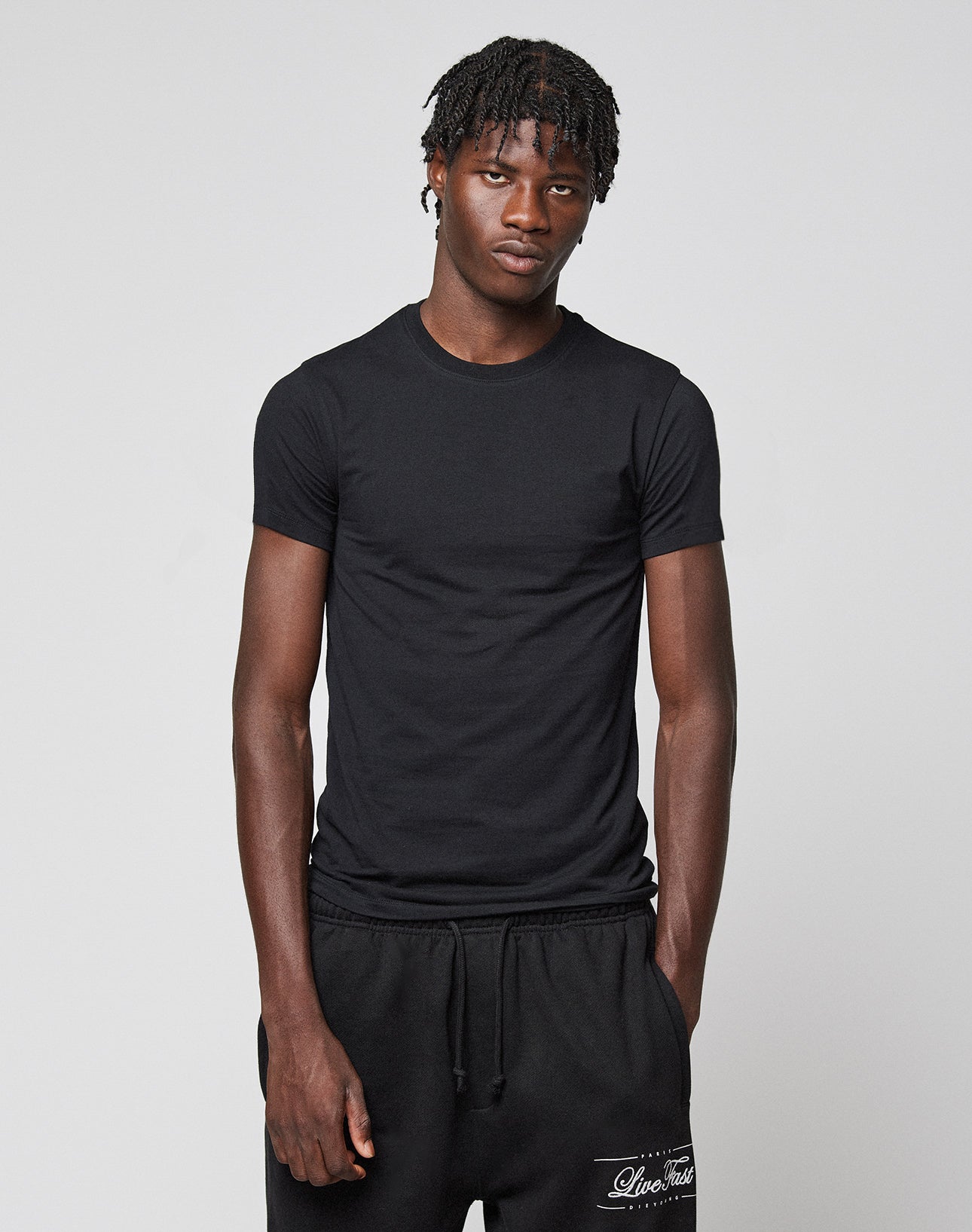A young man with short, textured hair wears the LIVE FAST DIE YOUNG LFDY Slim Tee and black sweatpants, standing against a plain light gray background with his hands by his sides.