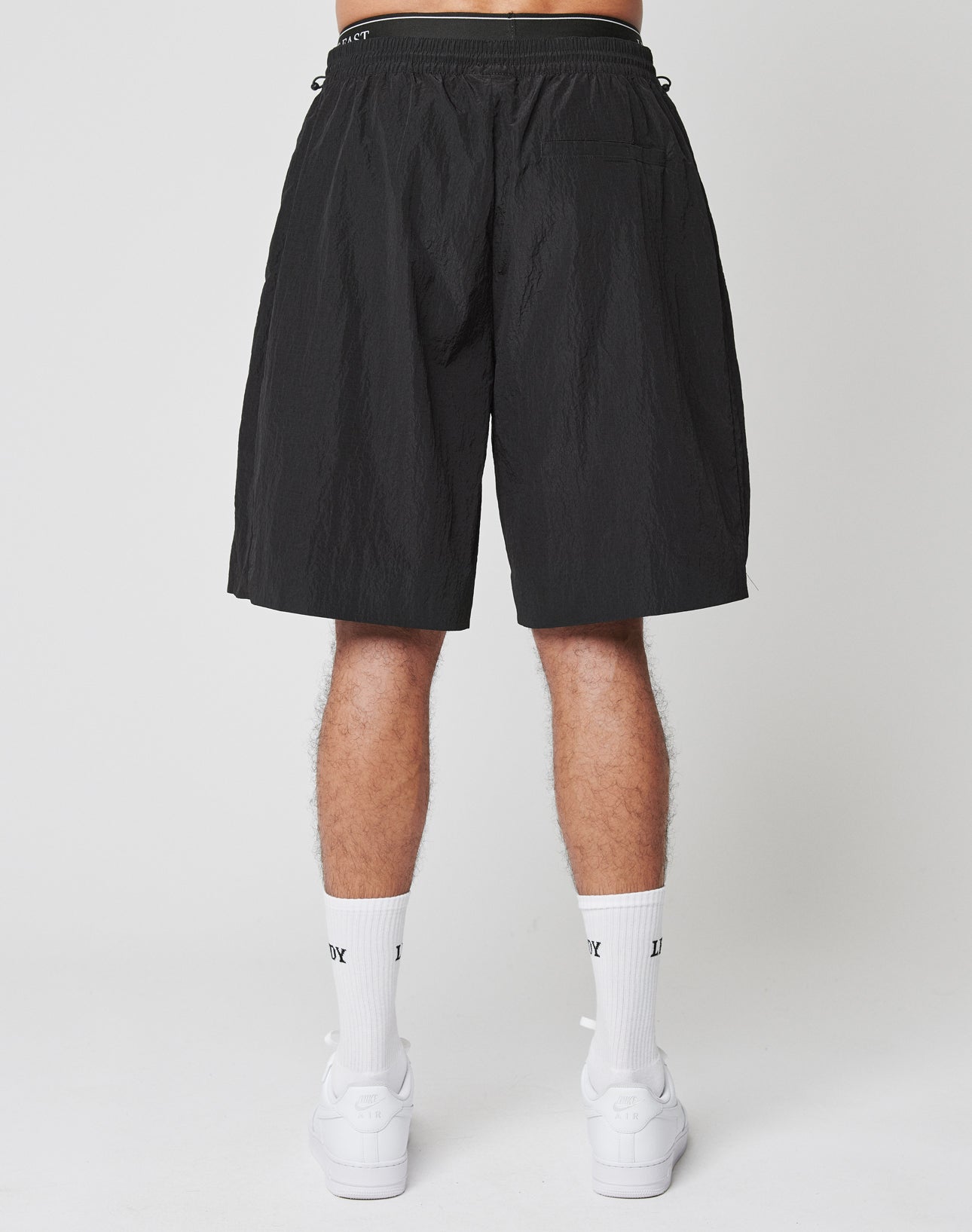 A person stands against a plain background, shown from the waist down, wearing LIVE FAST DIE YOUNG Live Fast Nylon Shorts with an oversized fit, white crew socks, and white sneakers. The shorts feature a single back pocket on the right side.