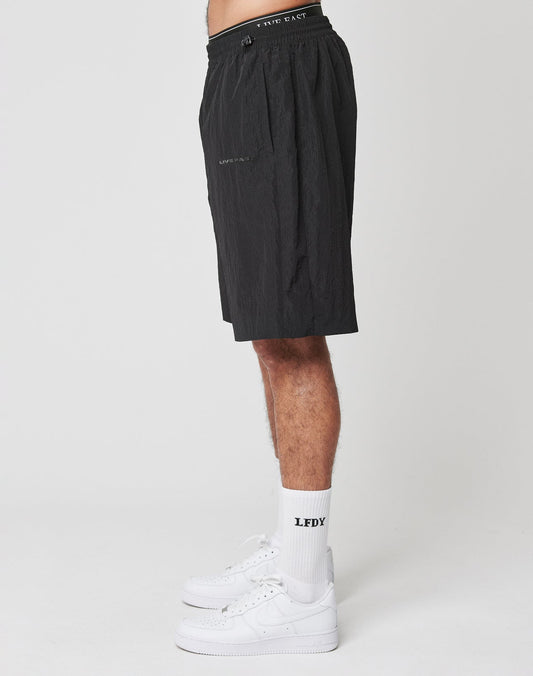 A person stands sideways against a white background, showing only the lower body, wearing black Live Fast Nylon Shorts from LIVE FAST DIE YOUNG, white LFDY socks, and white Nike sneakers.
