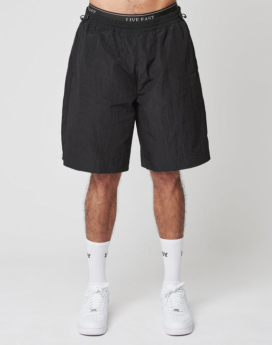 A person wears LIVE FAST DIE YOUNGs Live Fast Nylon Shorts, which feature an oversized fit and a bold Silikon-Print waistband, paired with white crew socks and sneakers. The setting is a plain light background; only the lower body is visible.