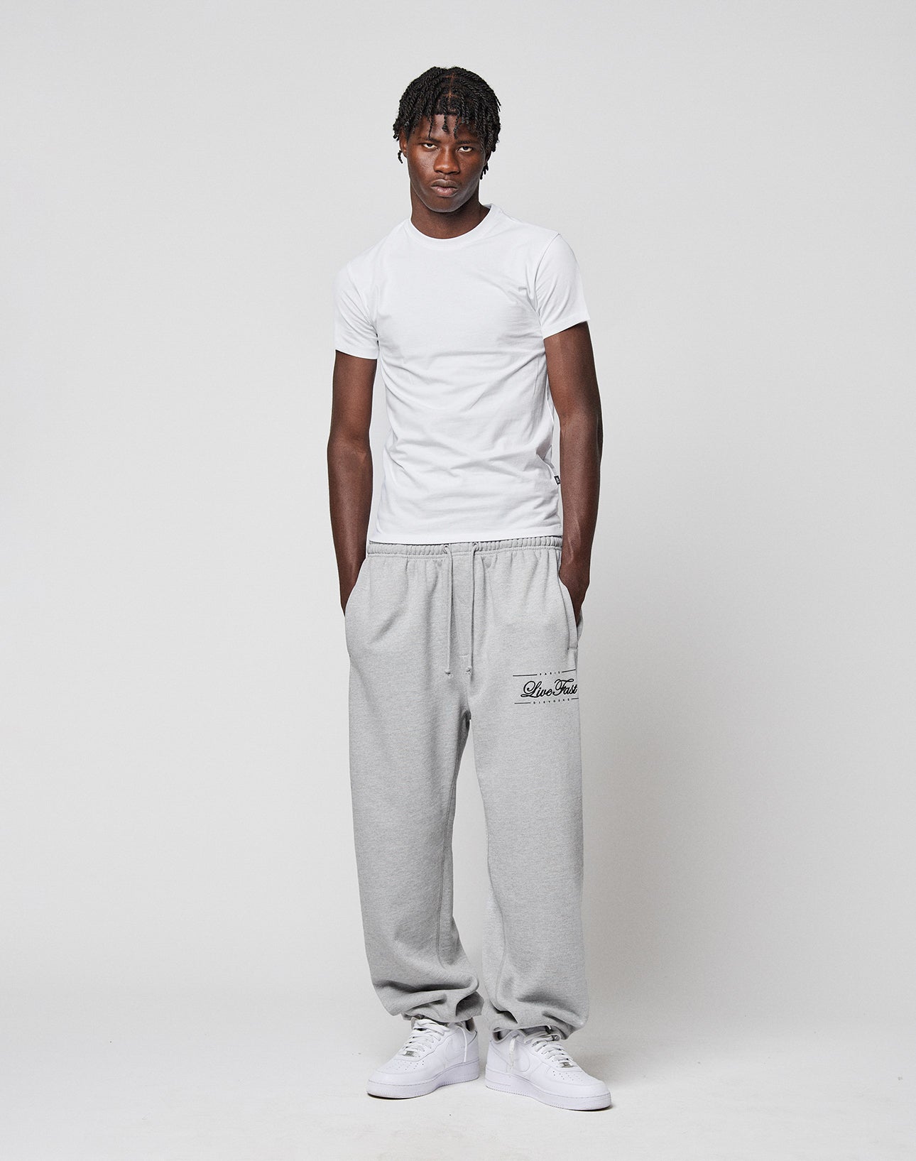A man stands against a plain background, wearing the LIVE FAST DIE YOUNG LFDY Slim Tee, loose gray sweatpants with "Dior Tears" and a flag label on the left leg, and white sneakers. His hands are in his pockets.