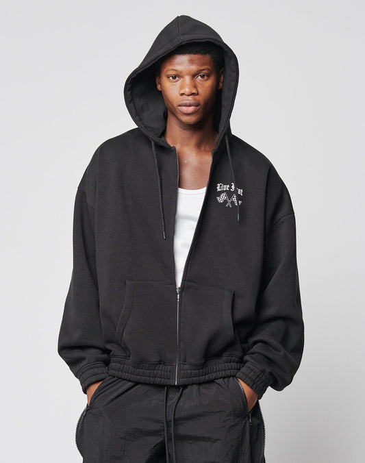 A person wears the LIVE FAST DIE YOUNG Ride or Die Hooded Zip in black with doppellagige Kapuze, a small white chest graphic, over a white tank top, plus black pants with elastischer Bund, standing against a plain light gray background.