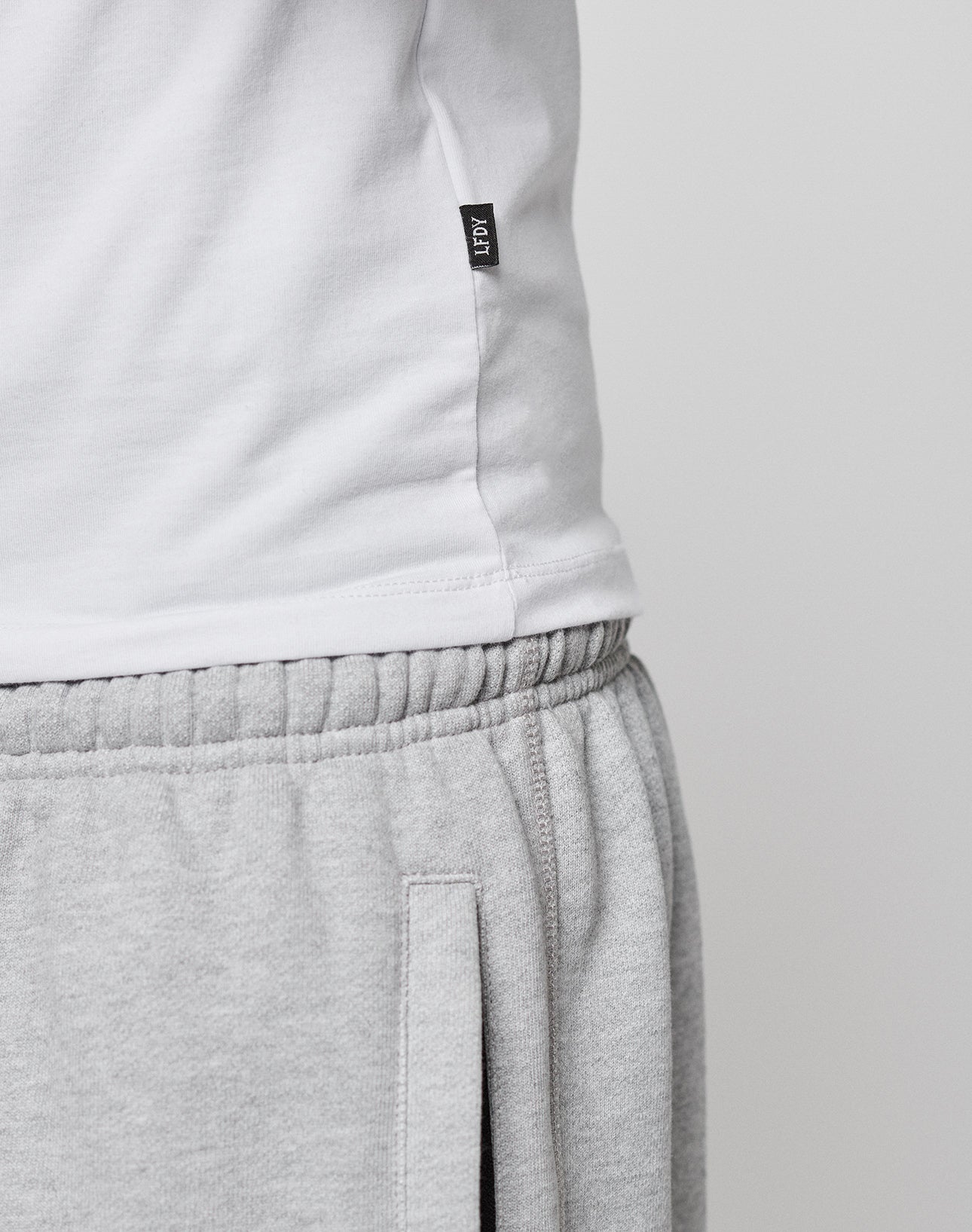 Close-up of a person wearing the white LFDY Slim Tee by LIVE FAST DIE YOUNG with a small black flag label, paired with light gray sweatpants featuring an elastic waistband and vertical pocket. The background is plain and light-colored.