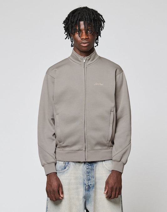 A young man with dark curly hair wears a LIVE FAST DIE YOUNG Sweatsuit Jacket in light gray and faded blue jeans, standing against a plain light gray background with a neutral expression.