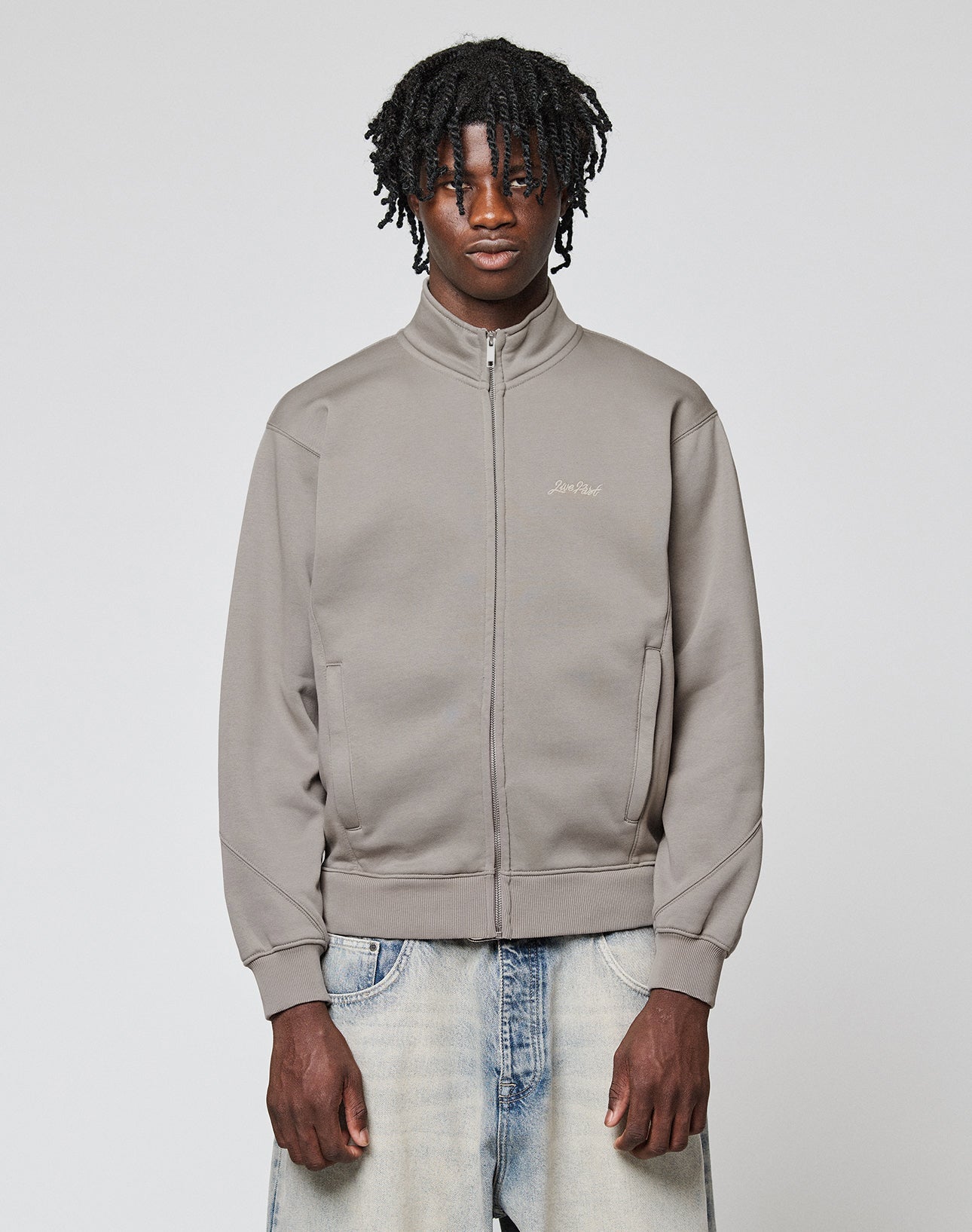 A young man with dark curly hair wears a LIVE FAST DIE YOUNG Sweatsuit Jacket in light gray and faded blue jeans, standing against a plain light gray background with a neutral expression.