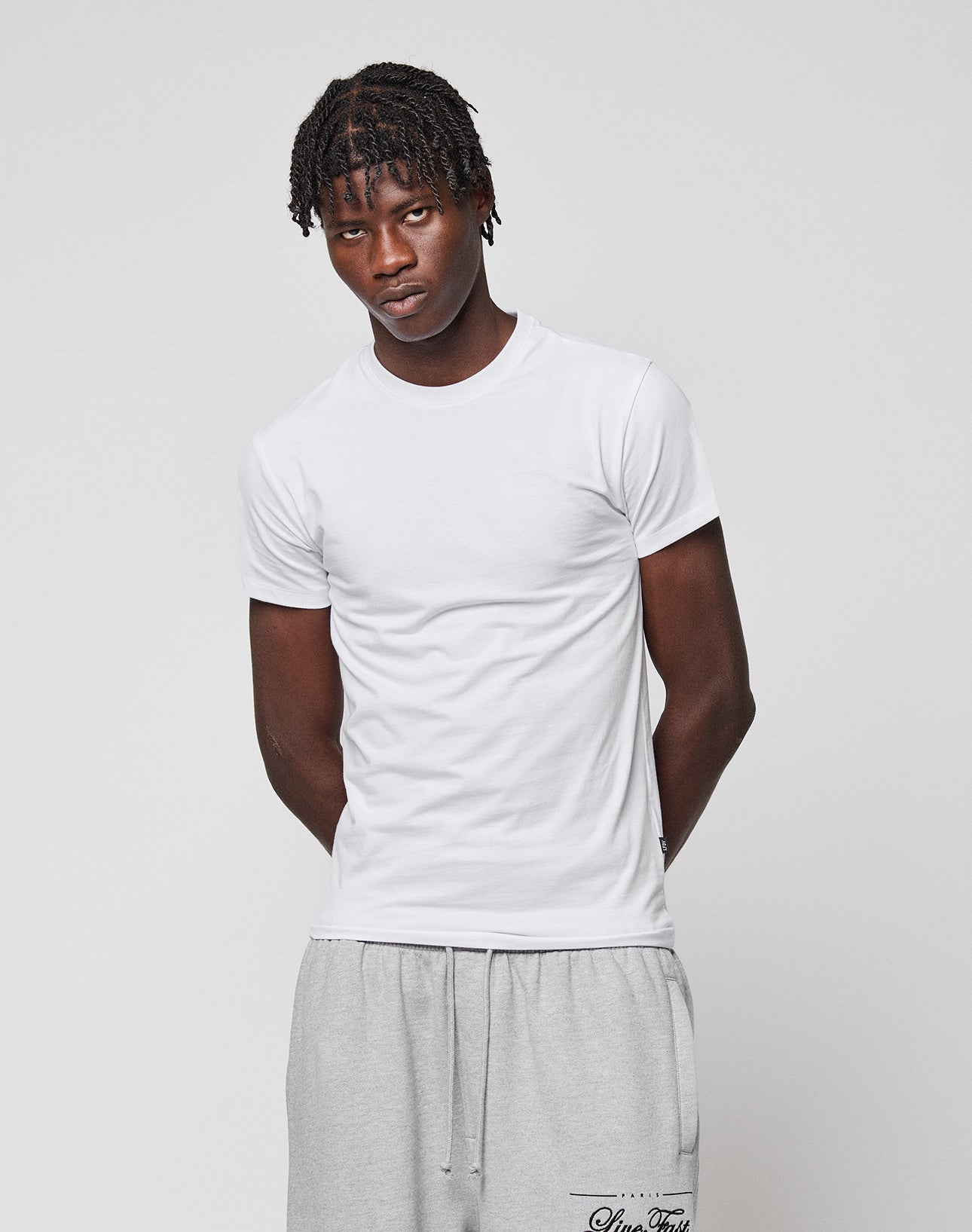 A person with short, twisted hair wears the LFDY Slim Tee by LIVE FAST DIE YOUNG and light gray drawstring sweatpants, standing against a plain light background with a neutral expression.