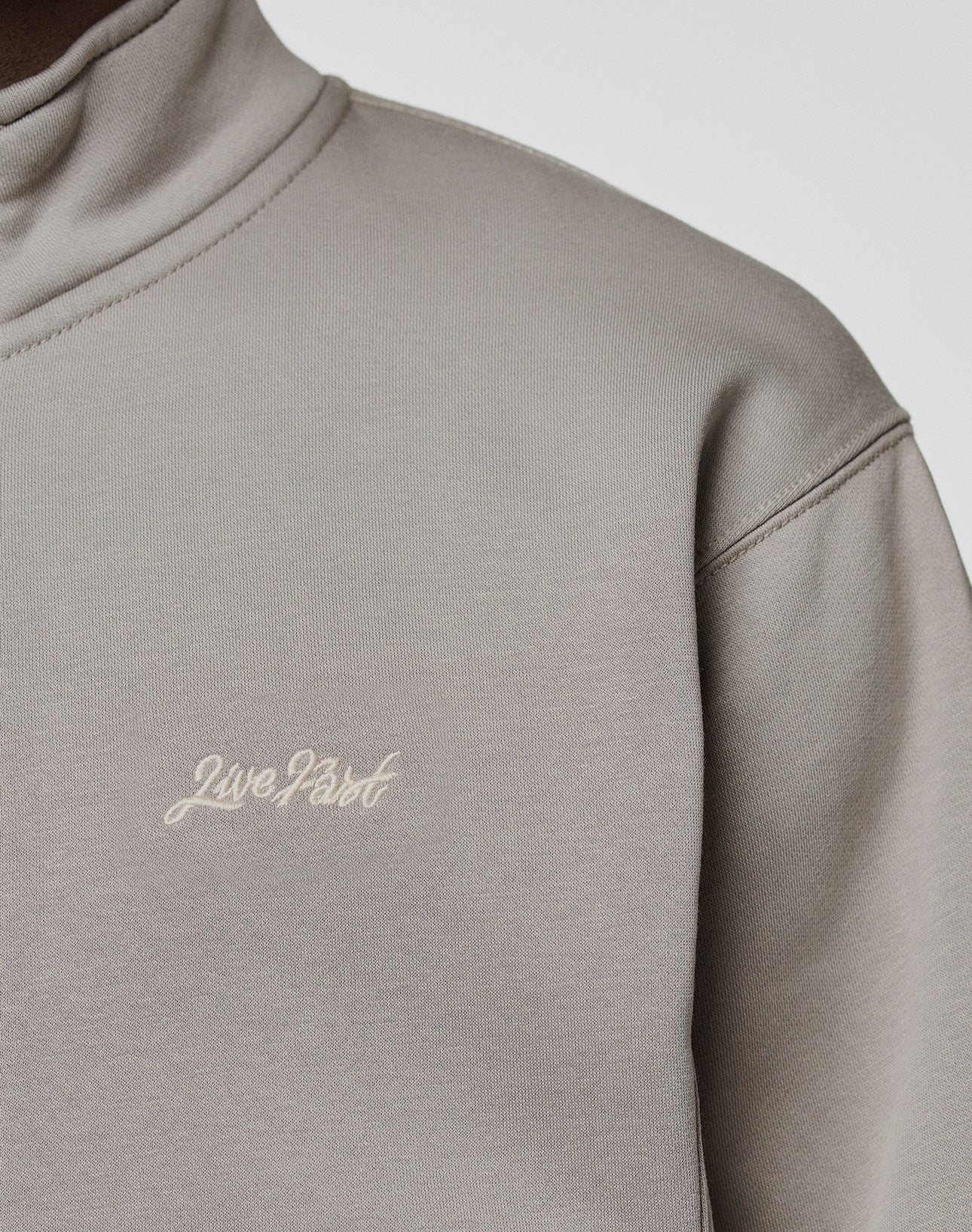 A close-up of a person wearing a light grey LIVE FAST DIE YOUNG Sweatsuit Jacket, Regular Fit, with "Live Fast" embroidered in small white cursive letters on the chest.