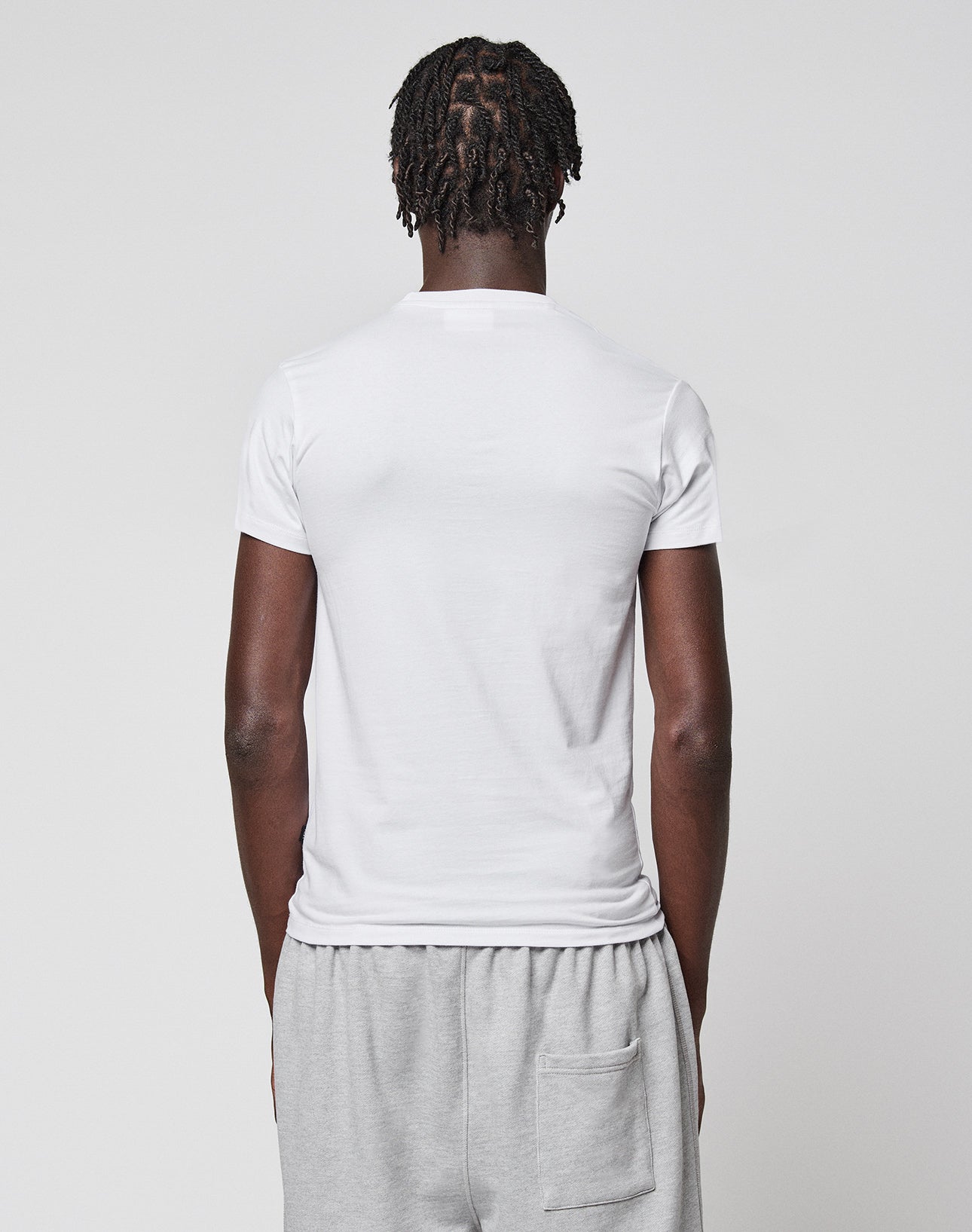 A person with short, twisted hair is seen from behind wearing a plain white LFDY Slim Tee by LIVE FAST DIE YOUNG and light gray shorts with a back pocket, standing against a plain light background.