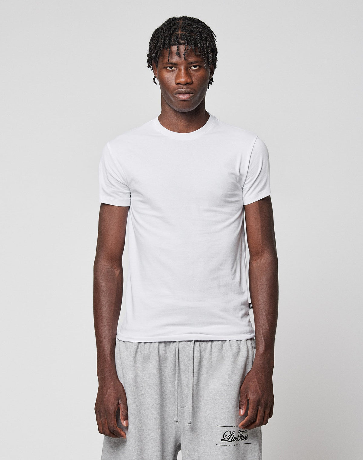 A young man with short, twisted hair wears the LIVE FAST DIE YOUNG LFDY Slim Tee in white and light gray sweatpants, standing against a plain light gray background and looking directly at the camera with a neutral expression. | model