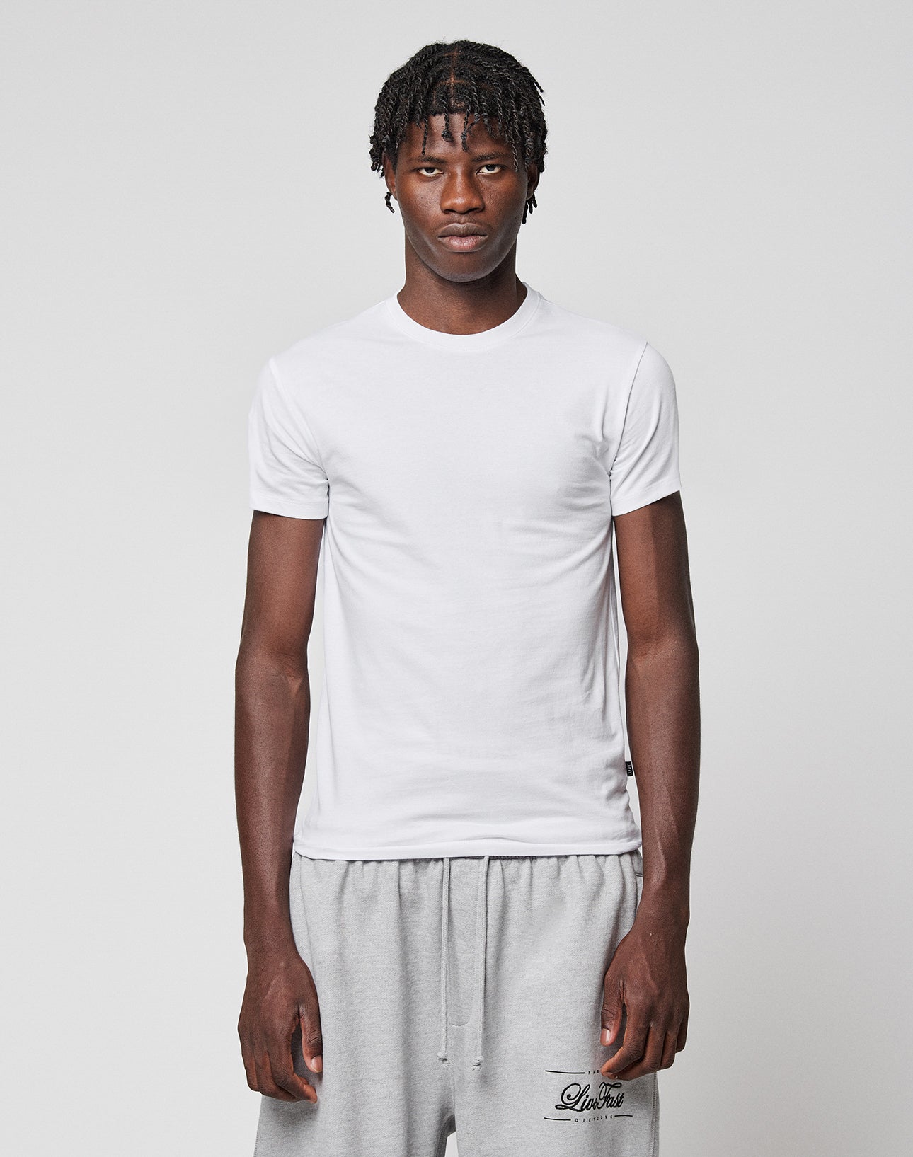 A young man with short, twisted hair wears the LIVE FAST DIE YOUNG LFDY Slim Tee in white and light gray sweatpants, standing against a plain light gray background and looking directly at the camera with a neutral expression.