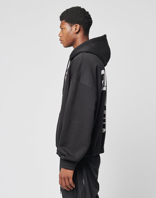 A young man stands in profile against a plain background, wearing the LIVE FAST DIE YOUNG Ride or Die Hooded Zip in black with an oversize fit, doppellagige Kapuze, black pants, and a bold white back design.