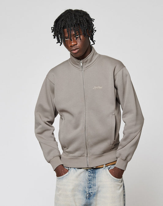 A young man with short dreadlocks wears the LIVE FAST DIE YOUNG Sweatsuit Jacket in Regular Fit with Stehkragen, hands in the pockets of light-washed jeans, standing against a plain, light background.