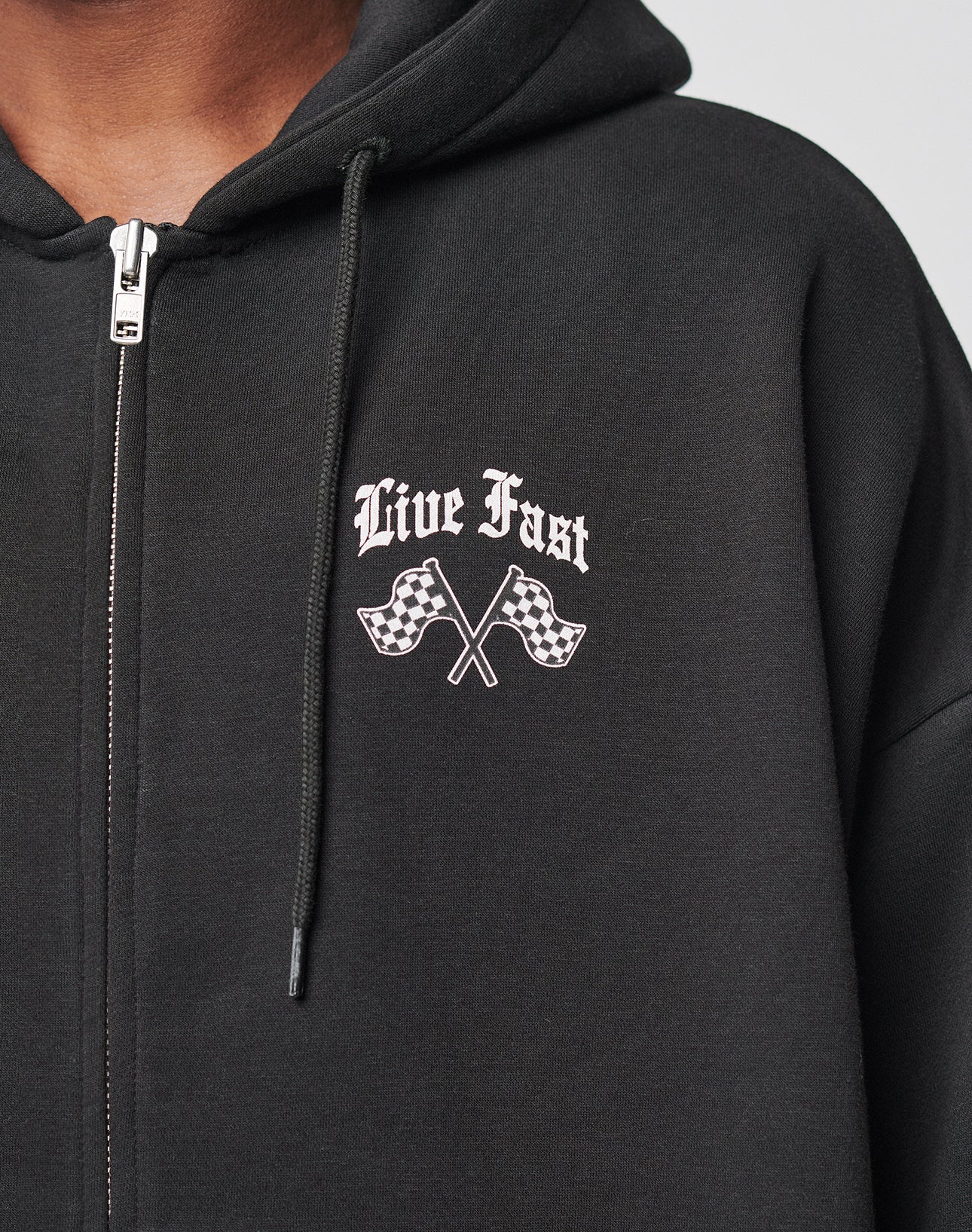 Someone wears the LIVE FAST DIE YOUNG Ride or Die Hooded Zip, a black oversized zip-up hoodie with Live Fast above two crossed checkered flags on the chest and a double-layered hood.