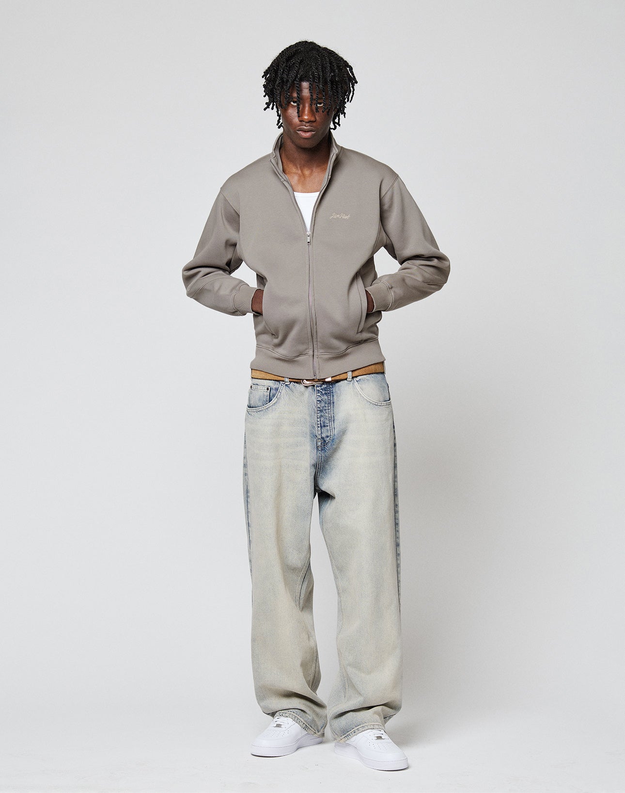 A man stands against a plain background wearing a LIVE FAST DIE YOUNG Sweatsuit Jacket in gray over a white shirt, Regular Fit light blue jeans, and white sneakers. He has his hands in his jacket pockets and looks at the camera.