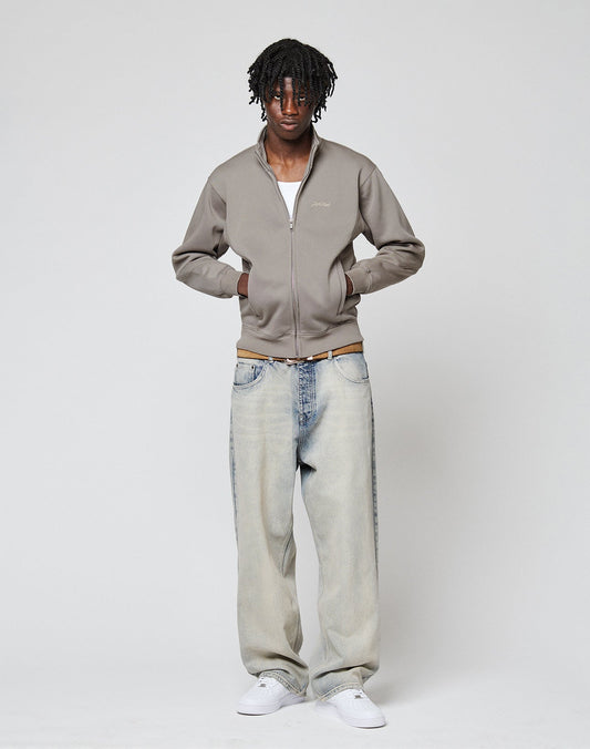 A man stands against a plain background wearing a LIVE FAST DIE YOUNG Sweatsuit Jacket in gray over a white shirt, Regular Fit light blue jeans, and white sneakers. He has his hands in his jacket pockets and looks at the camera.