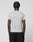 A person with short, twisted hair stands with their back to the camera, wearing a light gray LFDY Slim Tee by LIVE FAST DIE YOUNG and black pants. The tee features a subtle Flag-Label and is set against a plain light background.
