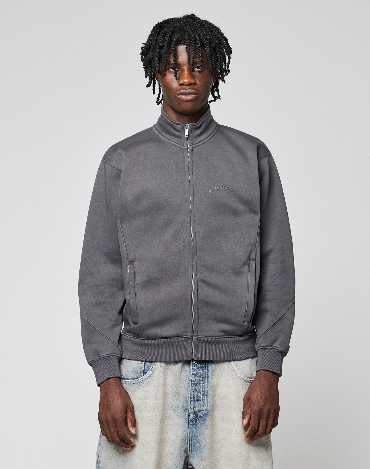 A young man with short dreadlocks wears a LIVE FAST DIE YOUNG Sweatsuit Jacket in dark gray and light blue jeans, standing against a plain light gray background and looking directly at the camera with a neutral expression.