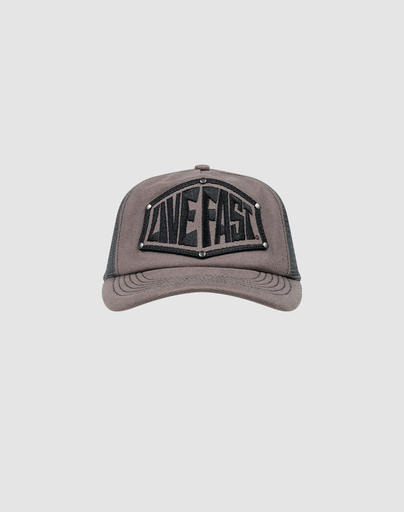 The LFDY Rivet Trucker by LIVE FAST DIE YOUNG is a gray cap with a black mesh back, LOVE FAST patch in bold black letters, silver stud accents, and a subtle flag-label detail.| plain 