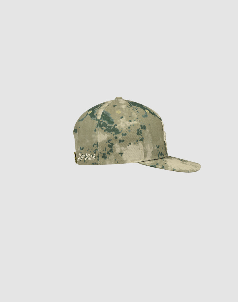 The LF Snapback by LIVE FAST DIE YOUNG is a green and beige camo baseball cap made from 100% Baumwolle, with a flat brim and white Logo-Stick text near the back. Model: LF2506-18254. Shown from the side on a light gray background.