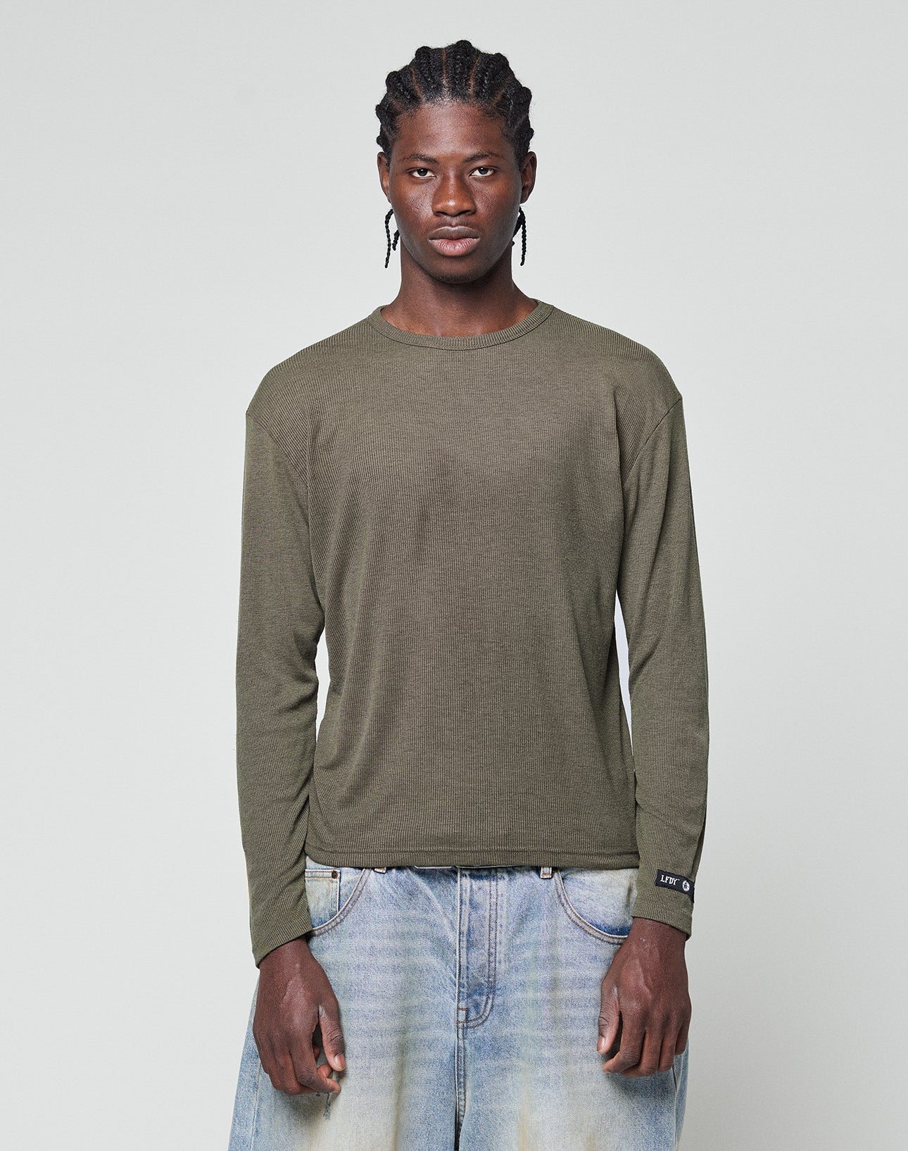 A person with braided hair wears an olive green LFDY Rib Longsleeve by LIVE FAST DIE YOUNG and light blue baggy jeans, standing against a plain light gray background. | model