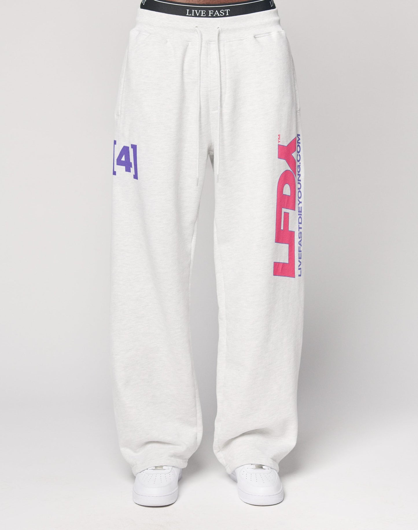 A person wears the LFDY Racing Wide Jogger by LIVE FAST DIE YOUNG, featuring large pink LFDY and purple text on the left leg, a wide elastic waistband with LIVE FAST, and pairs it with white sneakers against a plain white background.