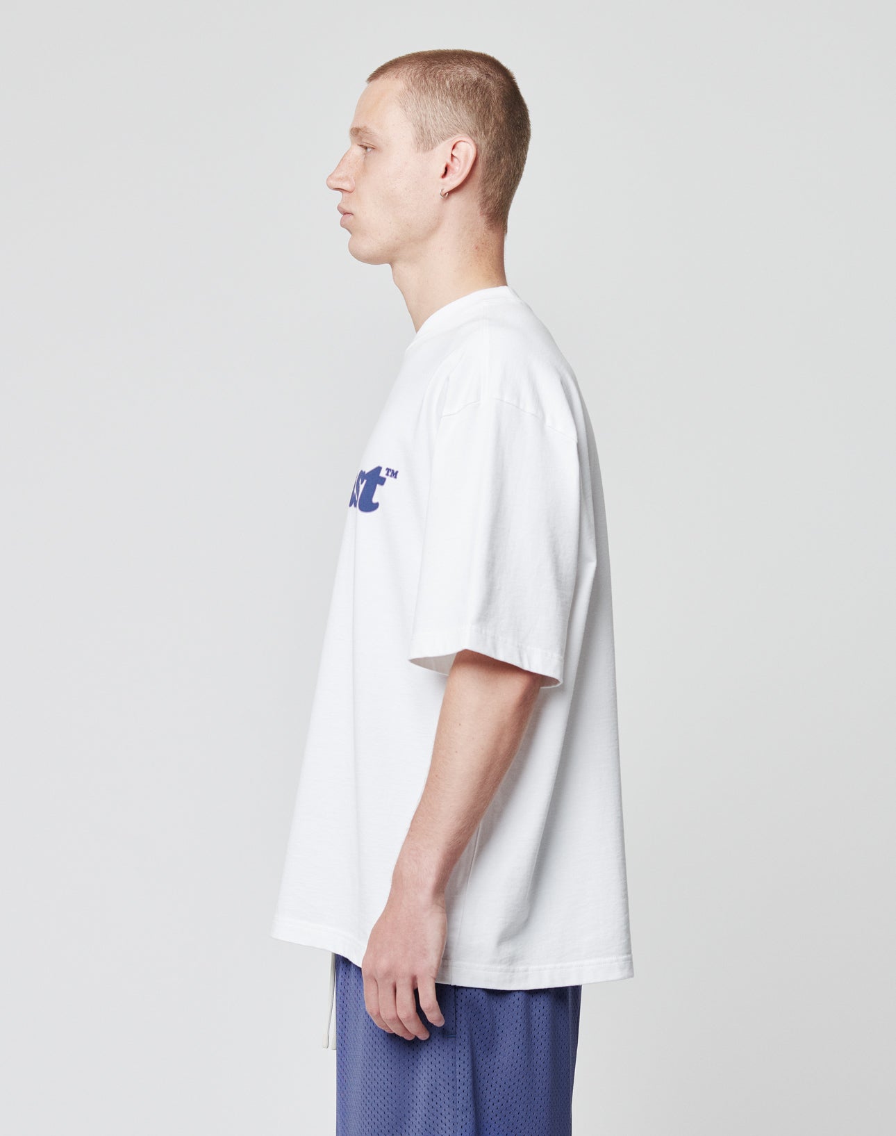 A young man with short hair stands in profile, wearing the LIVE FAST DIE YOUNG Basic Tee—an oversize fit white T-shirt made from 100% Baumwolle—with blue athletic shorts, set against a plain light gray background.